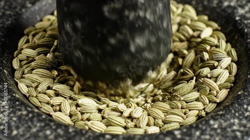 Close-up of green fennel seeds being crushed in a dark speckled granite mortar with a black pestle in natural light creating a natural textured background for culinary preparation and spice grinding