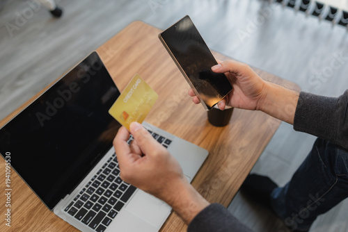 Man holding credit card and using smartphone near laptop for online payment and digital banking, representing e commerce transaction and modern financial technology.
