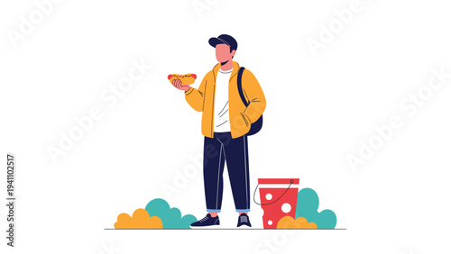 Young man standing outdoors wearing a yellow jacket and cap while holding a hotdog with a red bucket nearby him.