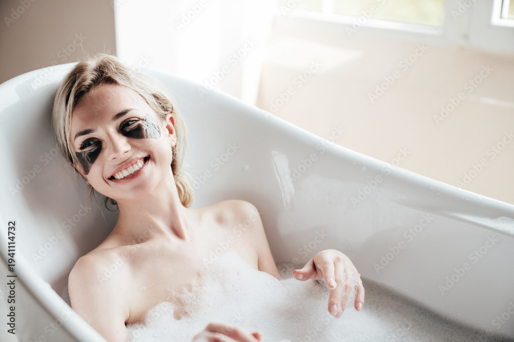 Obraz premium Portrait of young beautiful sexy woman having fun while lying in bathtub full of foam at home. Charming smiling model relaxing in luxury bath interior. Female enjoying beauty and skincare day