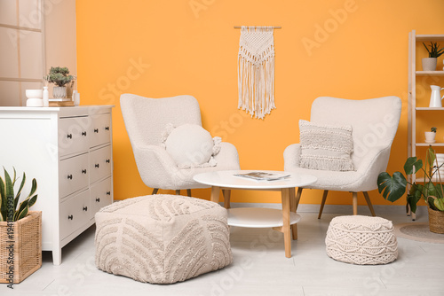 Interior of living room with table, poufs and soft armchairs near yellow wall