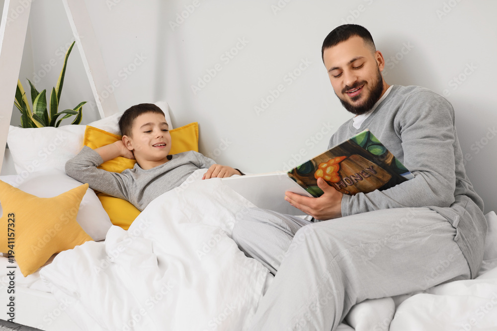 Fototapeta premium Happy father in pajamas reading book for his son on bed at home