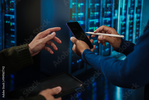 Close-up of hands using smartphone and stylus in data center, with server racks and blue lights in background, suggesting cybersecurity, IT support, device authentication and collaboration.