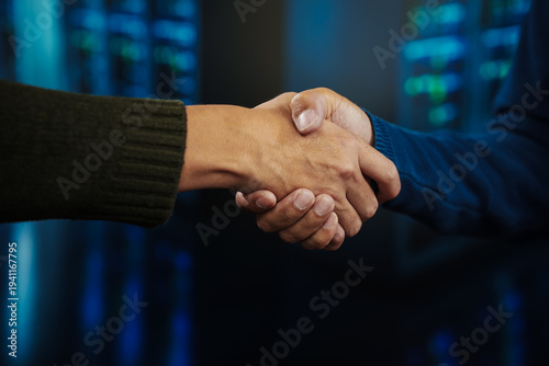 Close-up of two businesspeople shaking hands in a data center or server room with blue lights and blurred racks, symbolizing partnership, agreement, cybersecurity and technology collaboration.