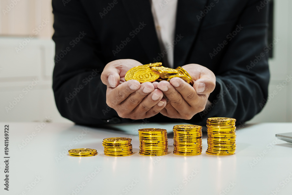 Fototapeta premium Businessman in suit holding gold coins in hands above stacks of coins on white desk, financial growth and savings concept, close-up with copy space.
