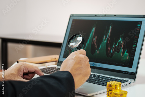 Investor uses magnifying glass to analyze stock candlestick chart on laptop, with stacked coins on desk, depicting financial research, trading strategy, and market analysis.