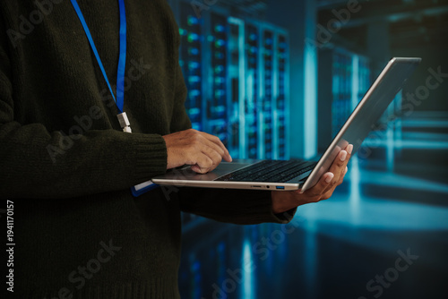 It professional using laptop in data center server room, cybersecurity and network admin concept with blue lights and racks in background
