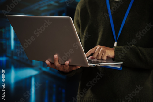 Close-up of professional using laptop in data center, blue network lights background, wearing lanyard badge; concept of cybersecurity, it administration, cloud computing, server maintenance.