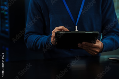 Close-up of IT technician with lanyard using a tablet in a server room, blue lighting, data center racks blurred in background; cybersecurity and system monitoring concept.