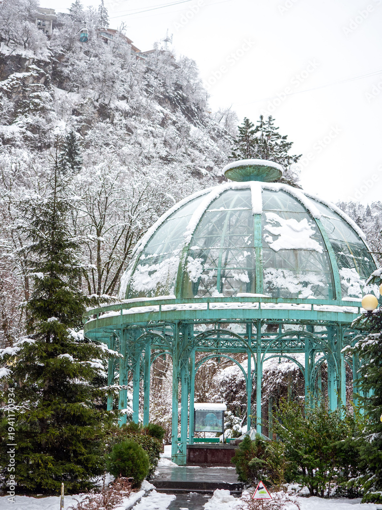 Fototapeta premium Winter scene of a glass gazebo in a snowy landscape with trees and a mountain background