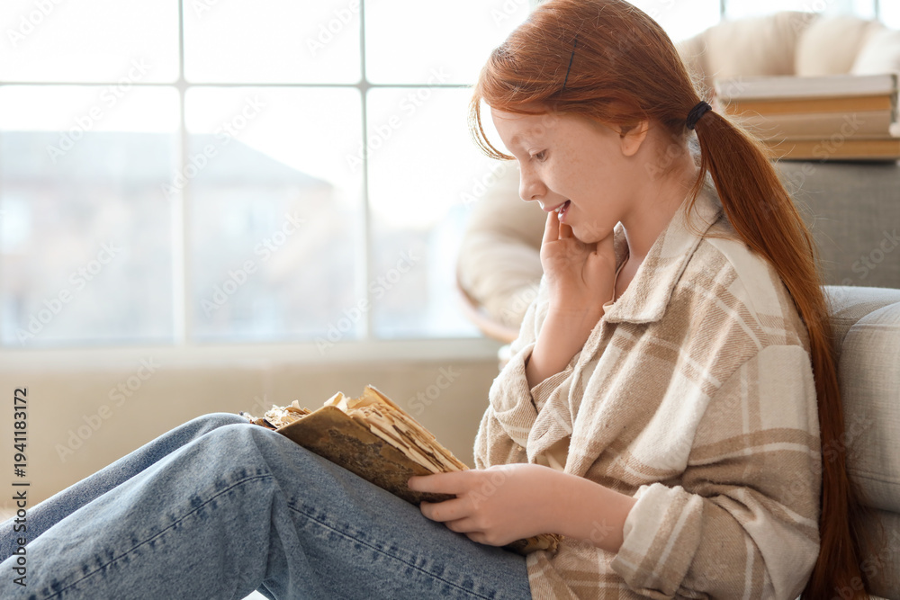 Fototapeta premium Cute teenage happy redhead girl with adventure book sitting on floor near window at home