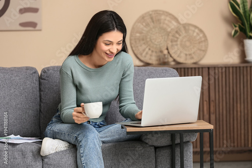 Tableau sur toile Beautiful young woman with cup of coffee and laptop sitting on sofa in living ro