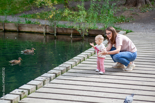 A mother and her young child are standing by a pond, watching ducks swim. The child is pointing at the ducks with excitement while the mother smiles and encourages.
