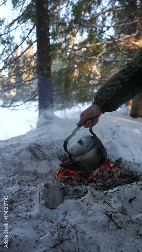 A hot drink from a vintage teapot is poured into a camping mug standing on the snow. Hot tea prepared over a campfire. Coffee is poured into a metal mug during a winter hike in the forest. The concept