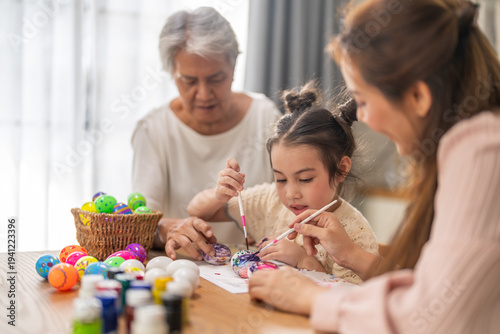 Asian little girl painting colorful easter eggs with brush while grandmother helping her at kitchen table, Three generations family spring holiday celebration, creative art craft learning, easter day