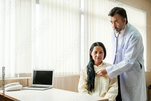 Φωτογραφία Doctor uses a stethoscope to examine a woman during a medical consultation in a clinic office, with a laptop and phone on the desk and blinds in the background