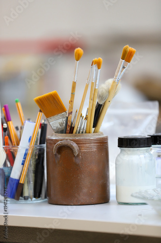 Pot en verre et pot en céramique contenant des crayons, stylo et pinceaux de différentes tailles. De petit pot de colle blanche à coté.