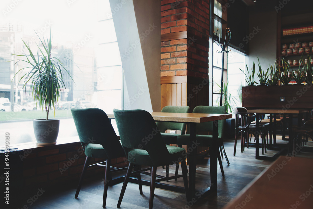 Fototapeta premium Inside a cafe, a table with four chairs sits next to a large window. Sunlight streams in. Plants decorate the space. More tables and chairs are visible in the background.