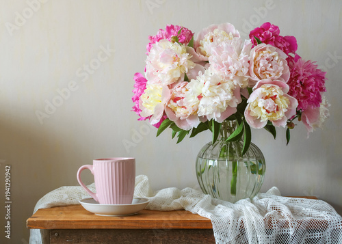 Bouquet of pink and white peonies in glass vase beside pastel cup on wooden table with soft window light cozy home interior romantic floral still life spring scene