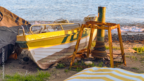 Photos A yellow and white boat sits on a sandy beach near the ocean at sunset A metal capstan structure and ropes are nearby in this coastal scene