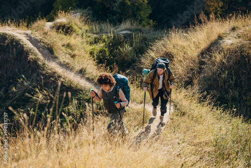 Fotografie Young family couple hikers walk uphill as use Nordic walking sticks