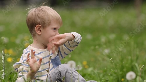 A fair-haired preschooler in a striped long-sleeve shirt sitting in a green meadow with dandelions, smiling and waving his hand. Medium shot, natural daylight, soft bokeh.