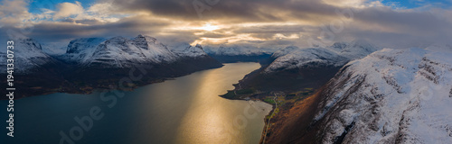 Aerial view of golden sunlight piercing through the clouds, reflecting off the still, dark waters amidst snow-capped mountains, Skibotn, Troms, Norway.