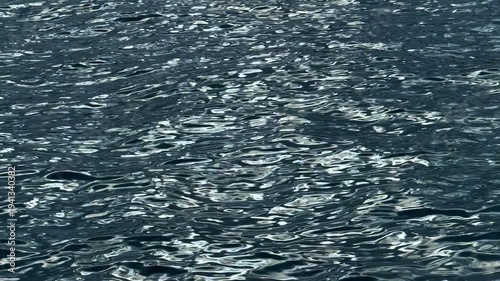 Rippled dark blue water surface with silver sunlight reflections. High angle view of textured water ripples with shimmering light glints and deep shadows.