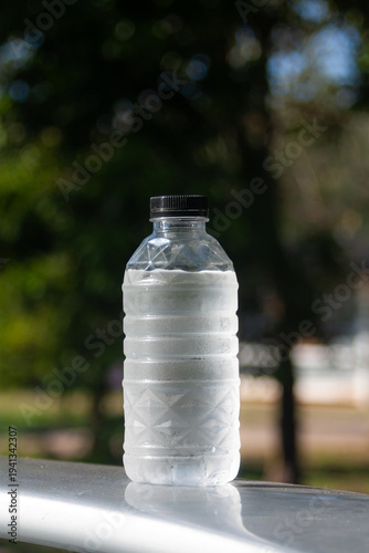 A cold plastic water bottle with condensation stands outdoors, catching sunlight against green blur.
