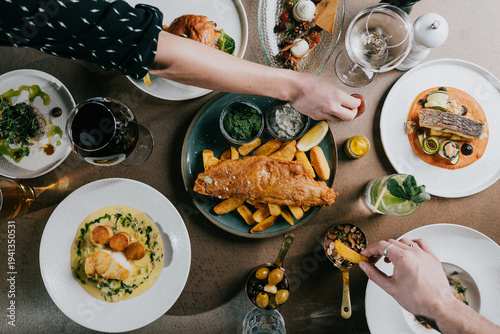 Overhead view of gourmet meal with fish, chips, and colorful side dishes. Hands reach for food, showing casual dining enjoyment. Plate arrangement suggests fine dining or celebratory gathering