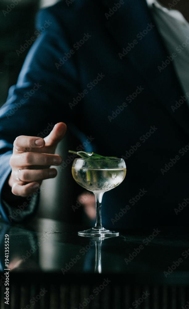 Obraz premium Bartender places a fresh mint leaf on a cocktail glass. Dark suit and gloved hand suggest elegance and precision. Glass holds clear liquid, possibly gin or champagne. Mood is sophisticated, calm