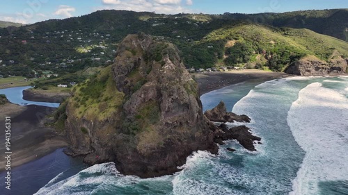 panning left from Lion Rock, a rocky headland to North Piha Beach on the western coast of the Auckland Region in New Zealand, while massive waves hitting the black sandy beach, 4k aerial video footag
