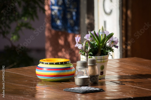 Ein Holztisch mit Tischdekoration bestehend aus einem Salz- und einem Pfefferstreuer, einem kleinen Blumentopf mit weissen Blüten, sowie einem bunten Aschenbecher mit farbigen Ringen