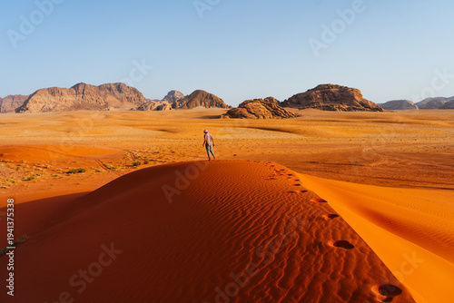 Traveler walking on a red sand dune in the Wadi Rum desert in Jordan. Scenic desert landscape with dramatic sandstone mountains and endless orange dunes under a clear blue sky. Famous Valley of the Mo