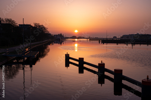 Impressive sunrise with intense colors reflected in the calm river, divided by wooden posts, the sunrise view from the dike bridge in 26382 Wilhelmshaven, Germany.