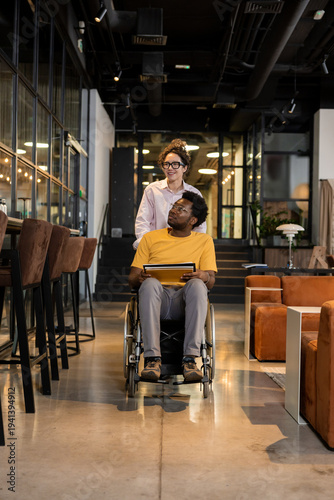 Female colleague assisting man in wheelchair moving through coworking office