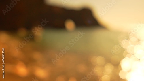 Bokeh sunset beach golden shimmering light reflecting off sea waves against blurred cliff background creating dreamy peaceful summer atmosphere
