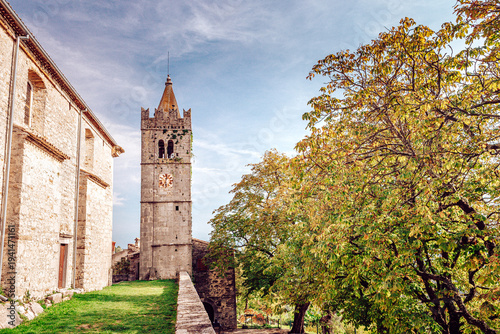 View of an old stone tower piercing the sky next to aged stone buildings and a colorful tree in Hum, Istria County, Croatia.