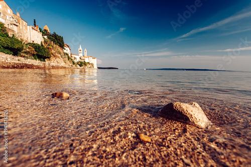 View of ancient stone buildings perched atop a cliff, reflecting in the clear, shallow waters lapping at the sandy shore, Rab, Primorje-Gorski Kotar County, Croatia.