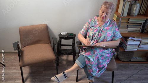 Senior elderly woman sits in a chair, draws and talks. Grandmother hobby.