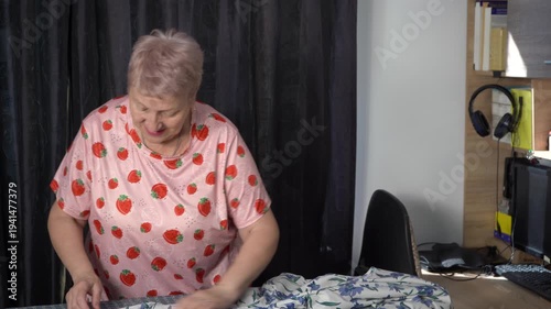 Elderly woman ironing clothes with an iron on a board. Grandmother chores routine