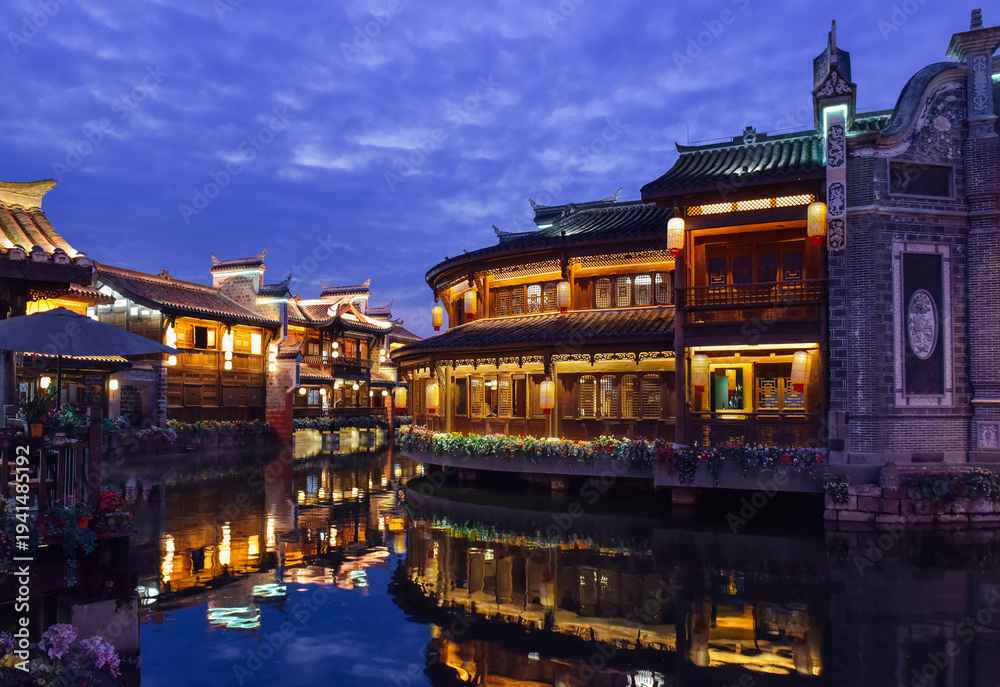 Obraz premium Night view of traditional stilt houses with glowing lanterns reflecting on the river in Moon Fields (Yueliangtian) scenic spot in Lizhuang Ancient Town, Yibin, Sichuan, China