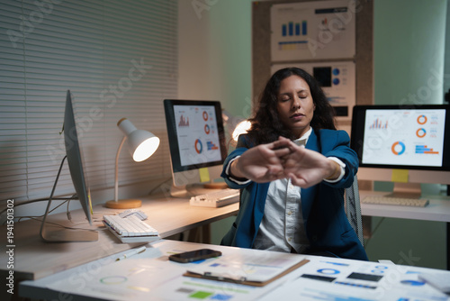 Businesswoman stretching at her desk in a dim office after overtime, rubbing tired shoulders amid computers and charts, capturing work-life imbalance and burnout risk