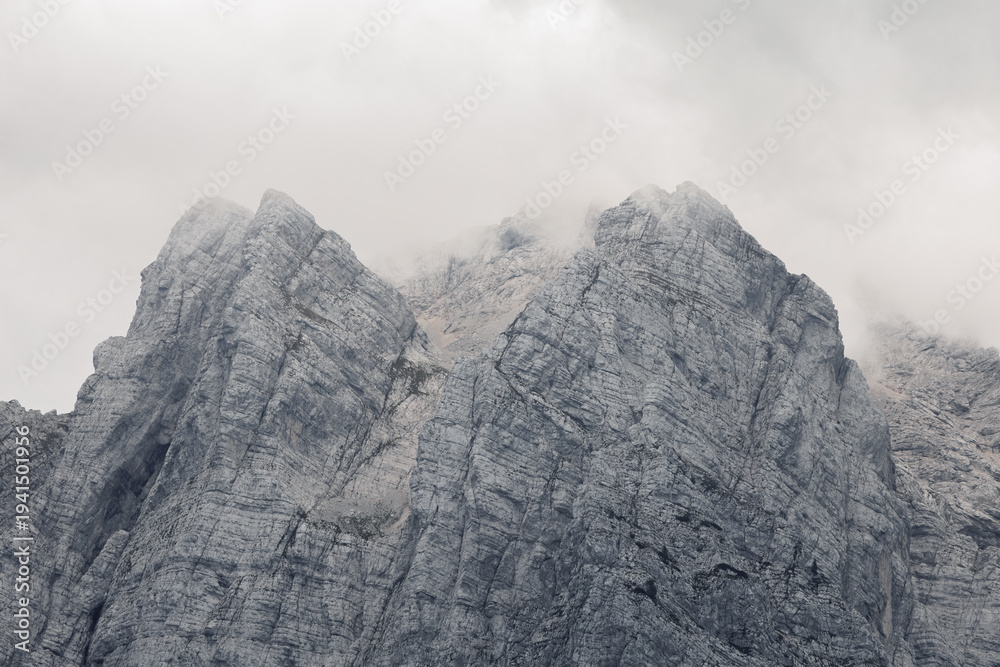 Fototapeta premium A somber, high-contrast view of jagged limestone mountain faces in the Julian Alps partially obscured by thick, low-hanging clouds on a misty day. Black and white textured stock photo