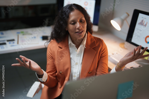 Businesswoman engaging in a video call and actively communicating, with multiple computer screens displaying data dashboards in an office environment during late hours