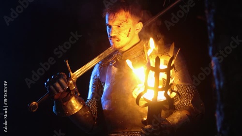 A knight in full armor, holding a sword and a torch, stands in a dark night forest. A medieval warrior with a bloodied face searches in the darkness.
