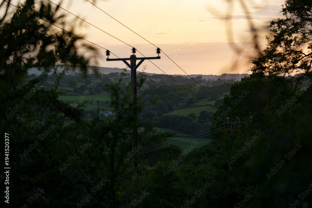 Obraz premium Rural landscape in Wales with power lines against a warm sunset. Golden light highlights rolling hills and countryside contours.