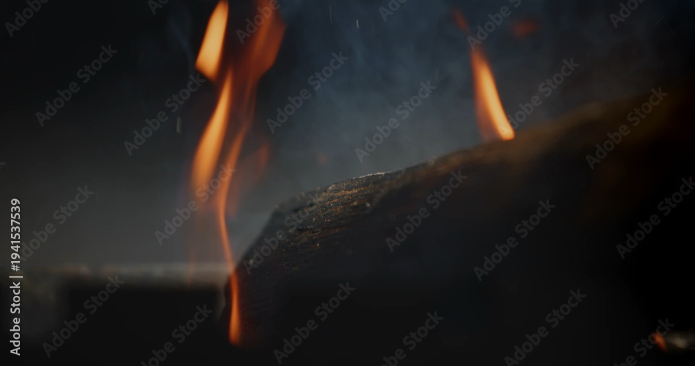 Fototapeta premium Low key close up of orange flames and smoke behind a dark log, creating a moody fire background for heat, danger, barbecue, and atmospheric concepts.