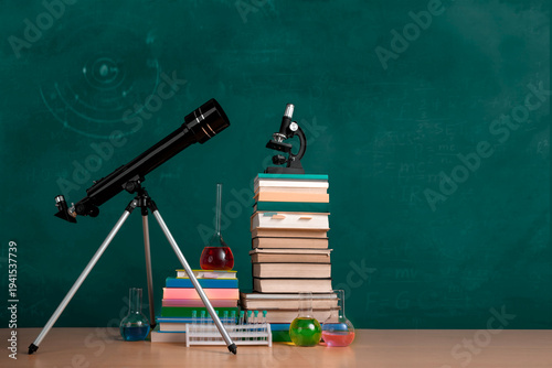 Natural sciences classroom scene with telescope, textbooks, microscope and chemistry glassware against green chalkboard. Blank space for text.