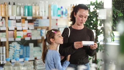 Woman in autumn clothes choosing baby ointment with her daughter near the counter in the pharmacy. High quality 4k footage